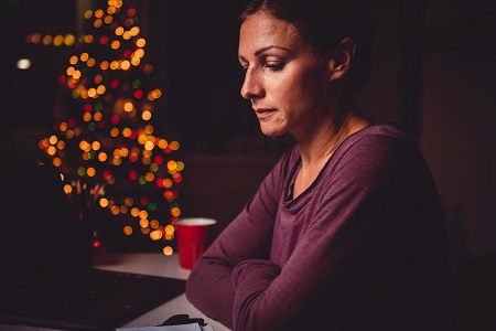 A woman is sitting at a counter at home, her arms folded with a phone in front of her. To the left is a blurred Christmas tree lit up with multi-coloured lights.